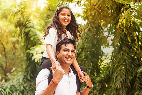 A man walking through a park with his daugher on his shoulders